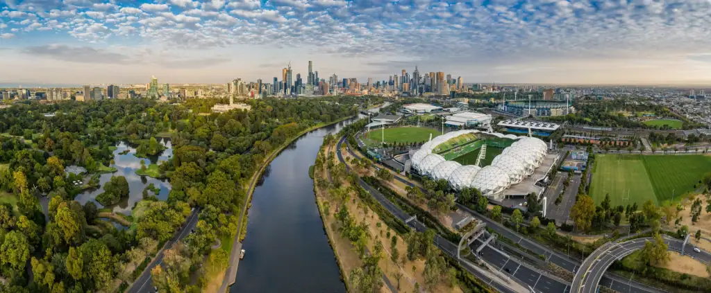 Aerial panoramic view of Melbourne CBD skyline with the Yarra River, Royal Botanic Gardens, AAMI Park, and Melbourne Park sports precinct under a cloudy sky